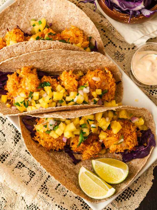 3 coconut shrimp tacos on a plate with pineapple salsa beside 2 lime slices, on a lace tablecloth.