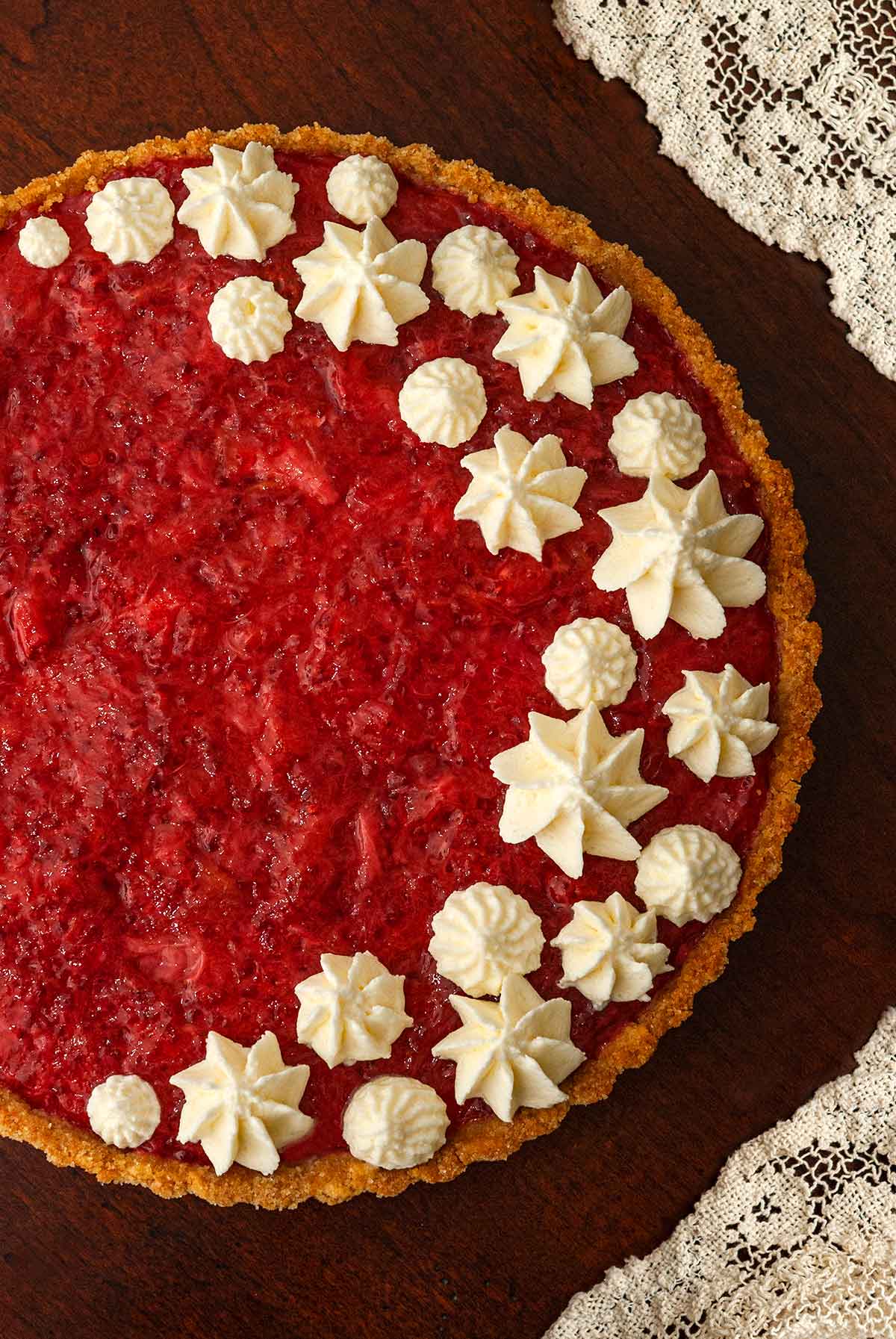 A strawberry rhubarb tart, topped with dots of whipped cream, beside a lace tablecloth.