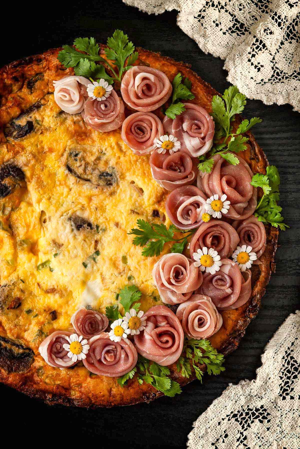 A quiche garnished with a wreath of prosciutto roses on a table beside a lace tablecloth.