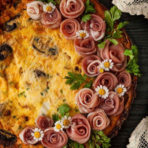 A quiche garnished with a wreath of prosciutto roses on a table beside a lace tablecloth.