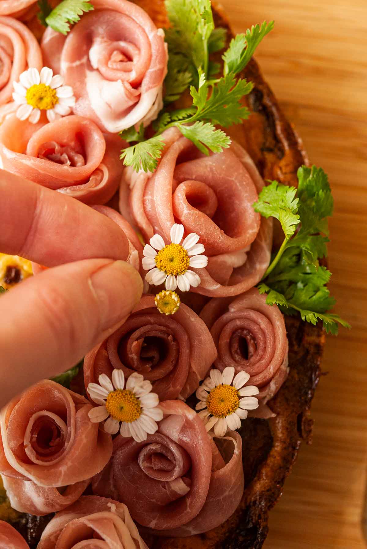 Fingers placing a small flower within a prosciutto rose wreath on a quiche.