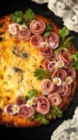 A quiche garnished with a wreath of prosciutto roses on a table beside a lace tablecloth.