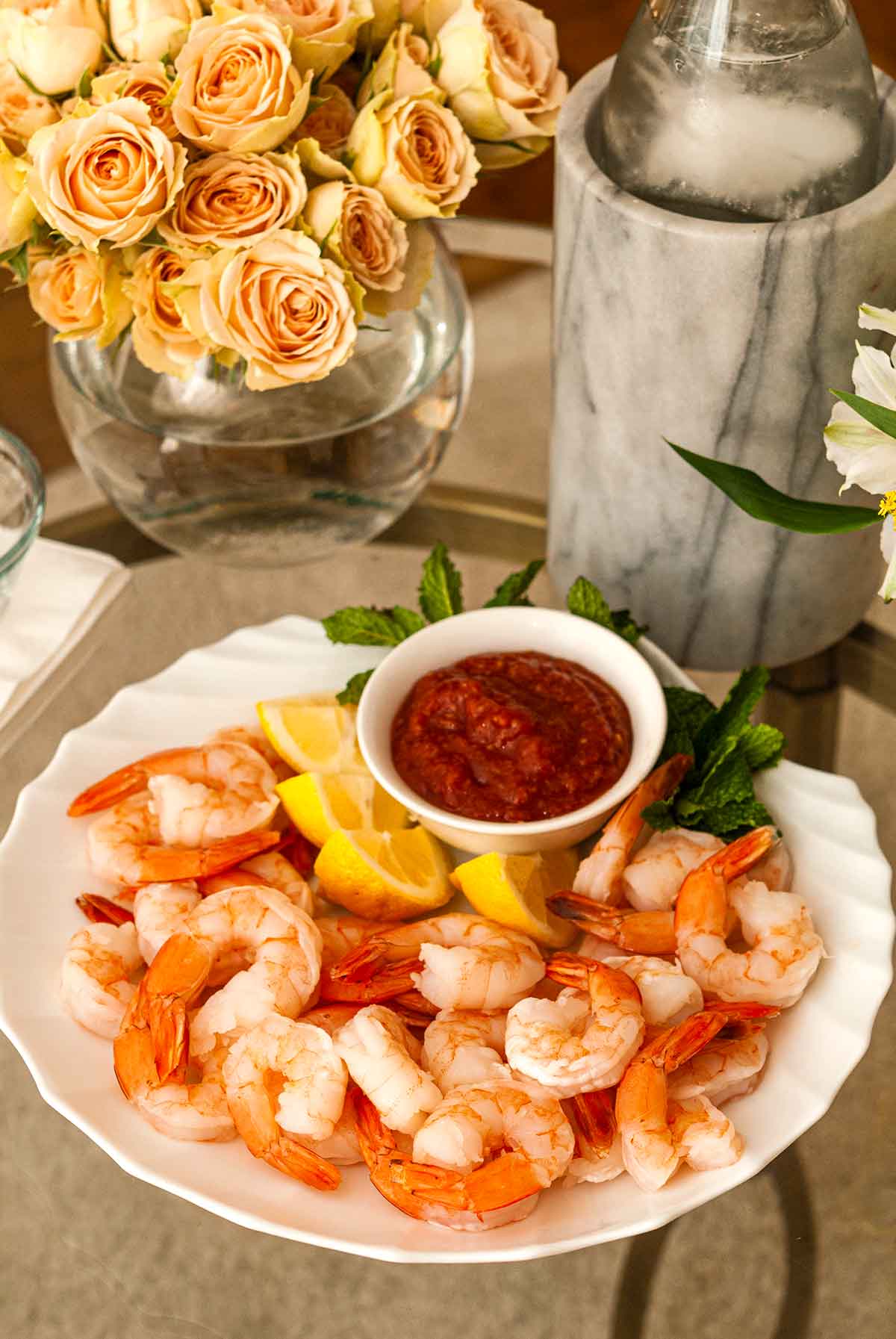 Shrimp cocktail on a shell-shaped plate beside roses and a bottle of water in a marble holder.