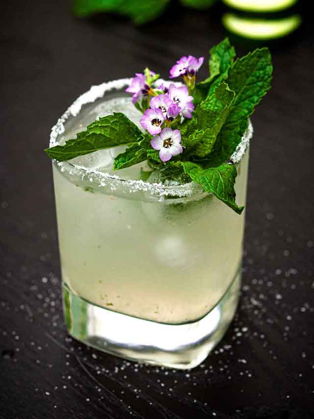 A cucumber mint margarita, garnished with small flowers and mint, on a table lightly sprinkled with salt.