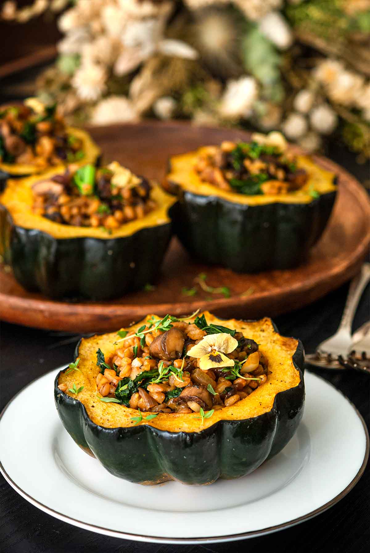1 stuffed acorn squash on a plate in front of 3 others in front of dry flowers on a table.