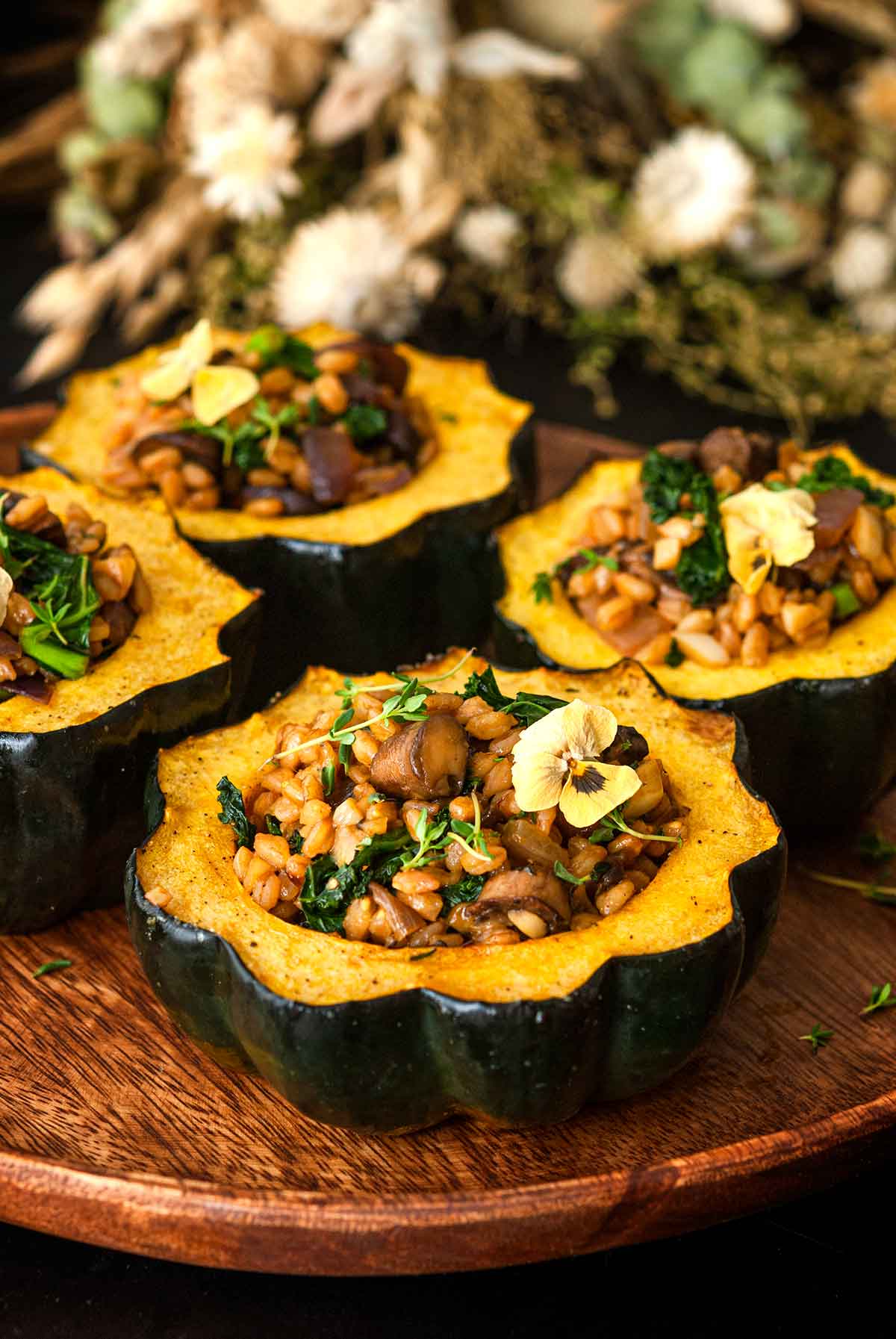 4 acorn squash on a wooden plate in front of dry flowers on a table.