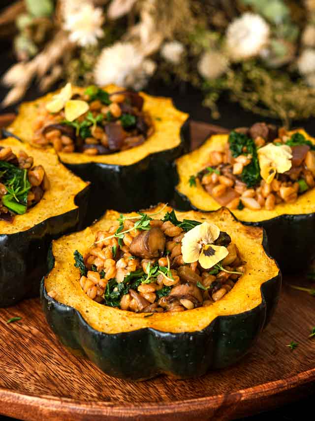 4 acorn squash on a wooden plate in front of dry flowers on a table.
