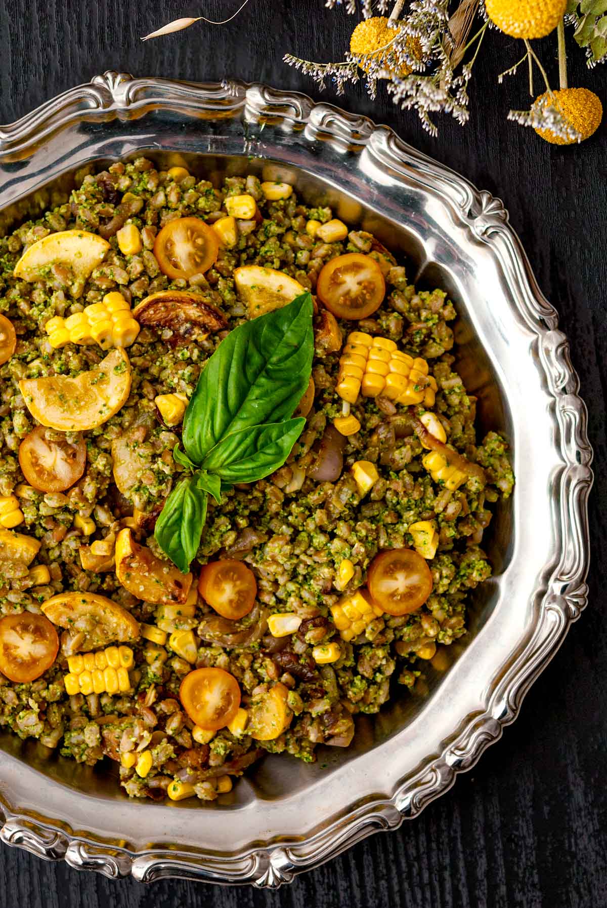 A decorative silver bowl with pesto farro and mixed vegetables on a table beside dry flowers..