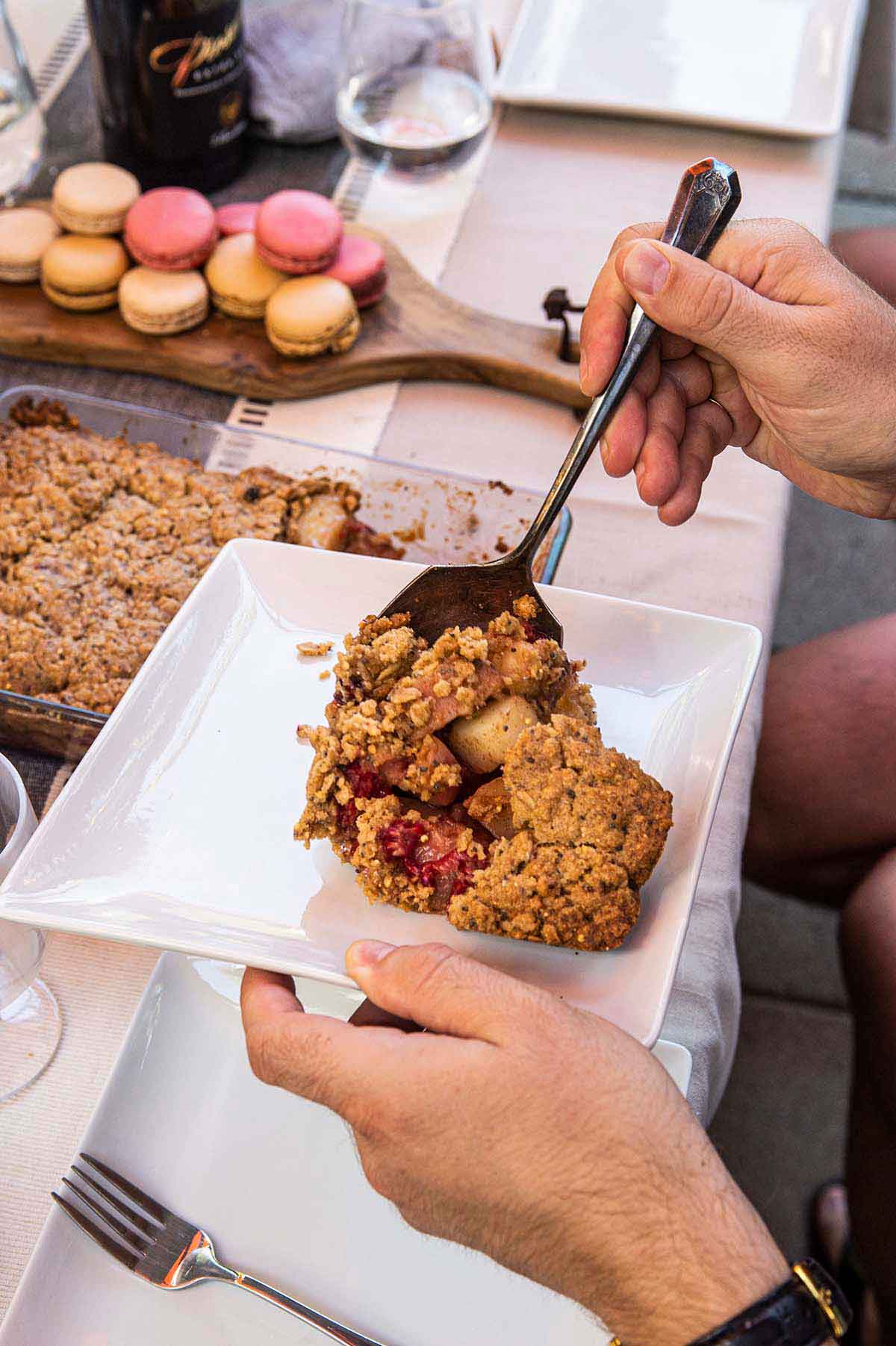 Hands serving peach crisp onto a plate with macrons in the background on a table.