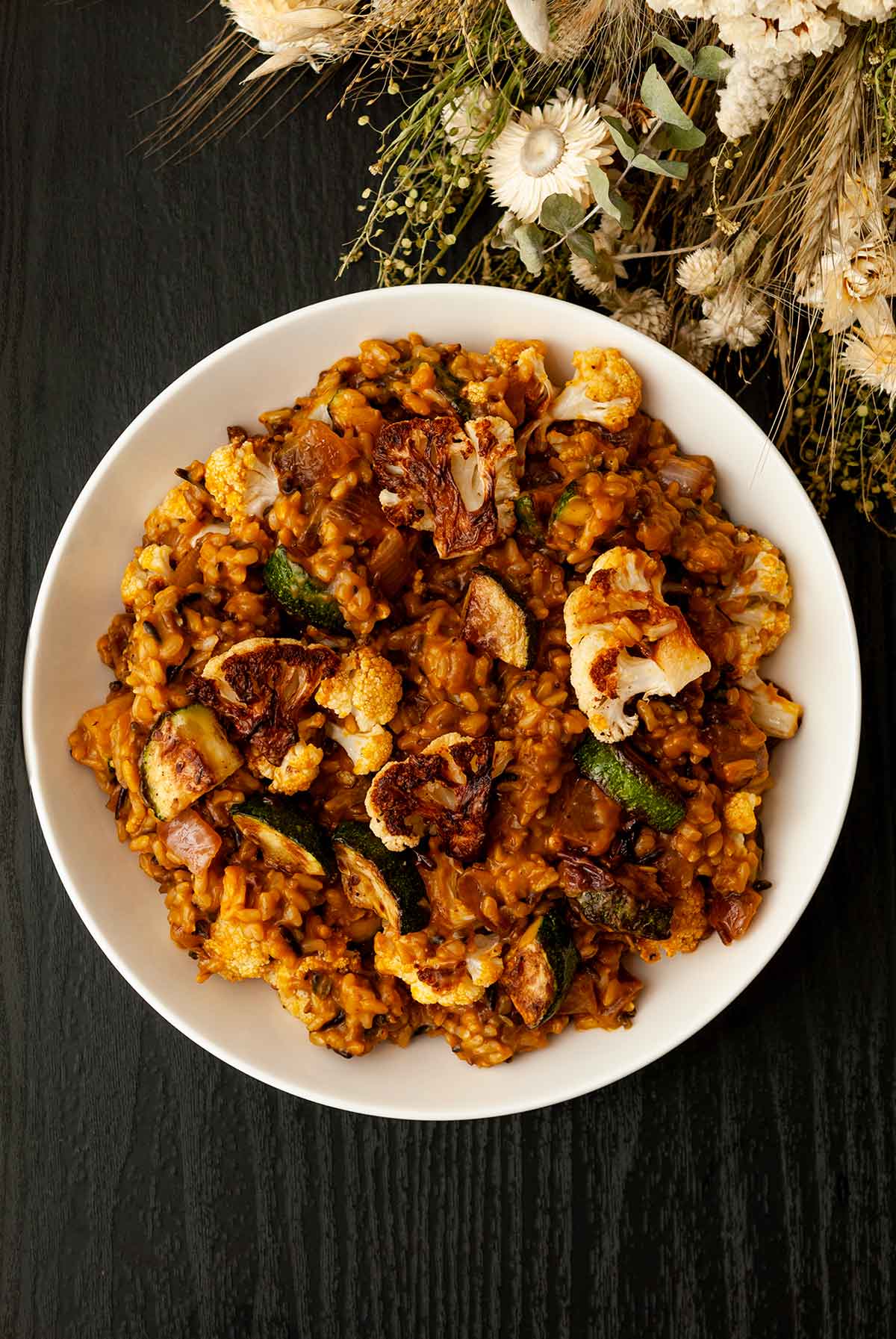 A centered bowl of pumpkin rice with fall vegetables on a table with dry flowers.