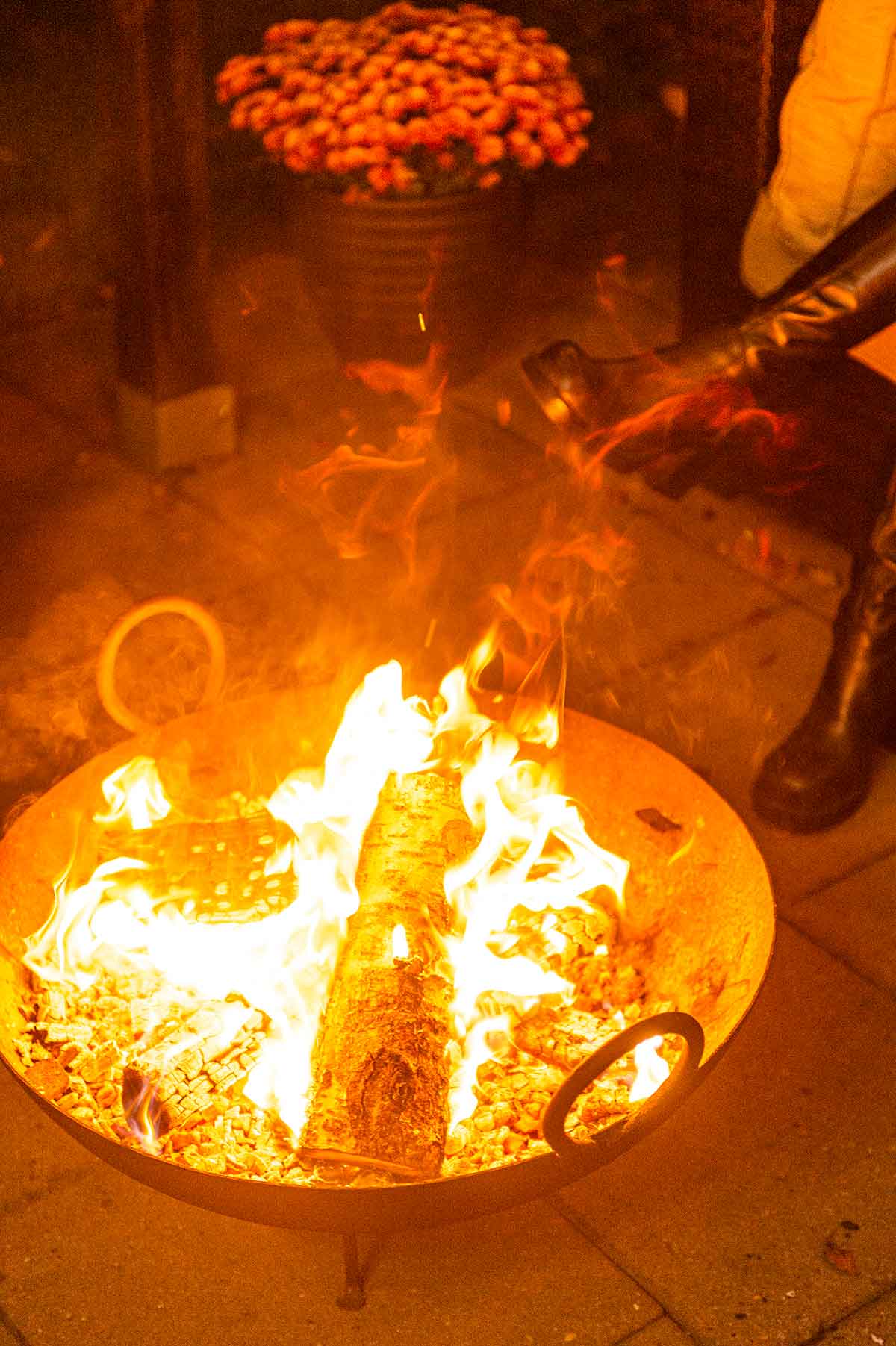 A firepit with fire and wood in front of a pot of mums and a person's boot.