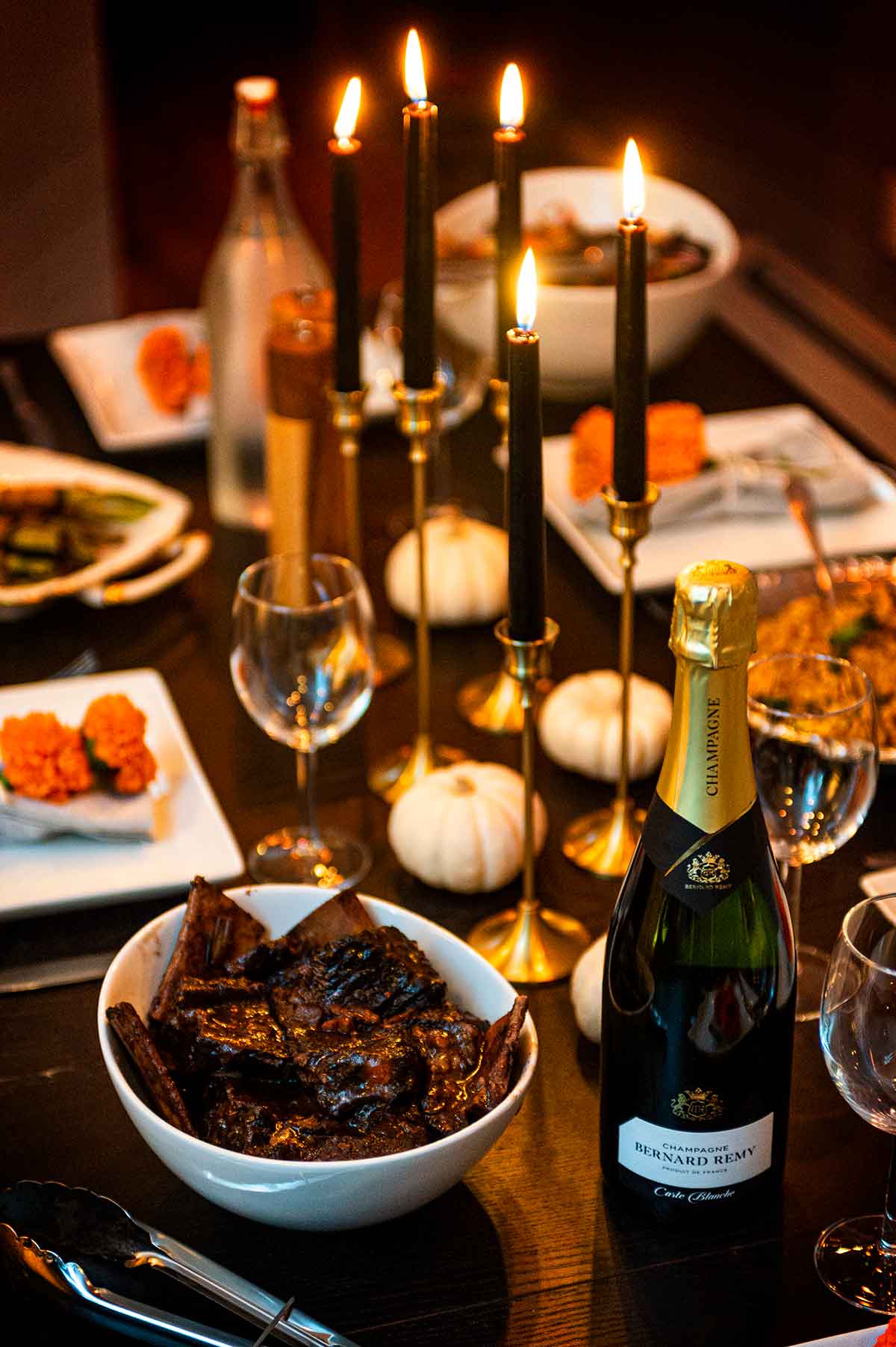 A dark dinner table with black candles, marigolds, short ribs and champagne.