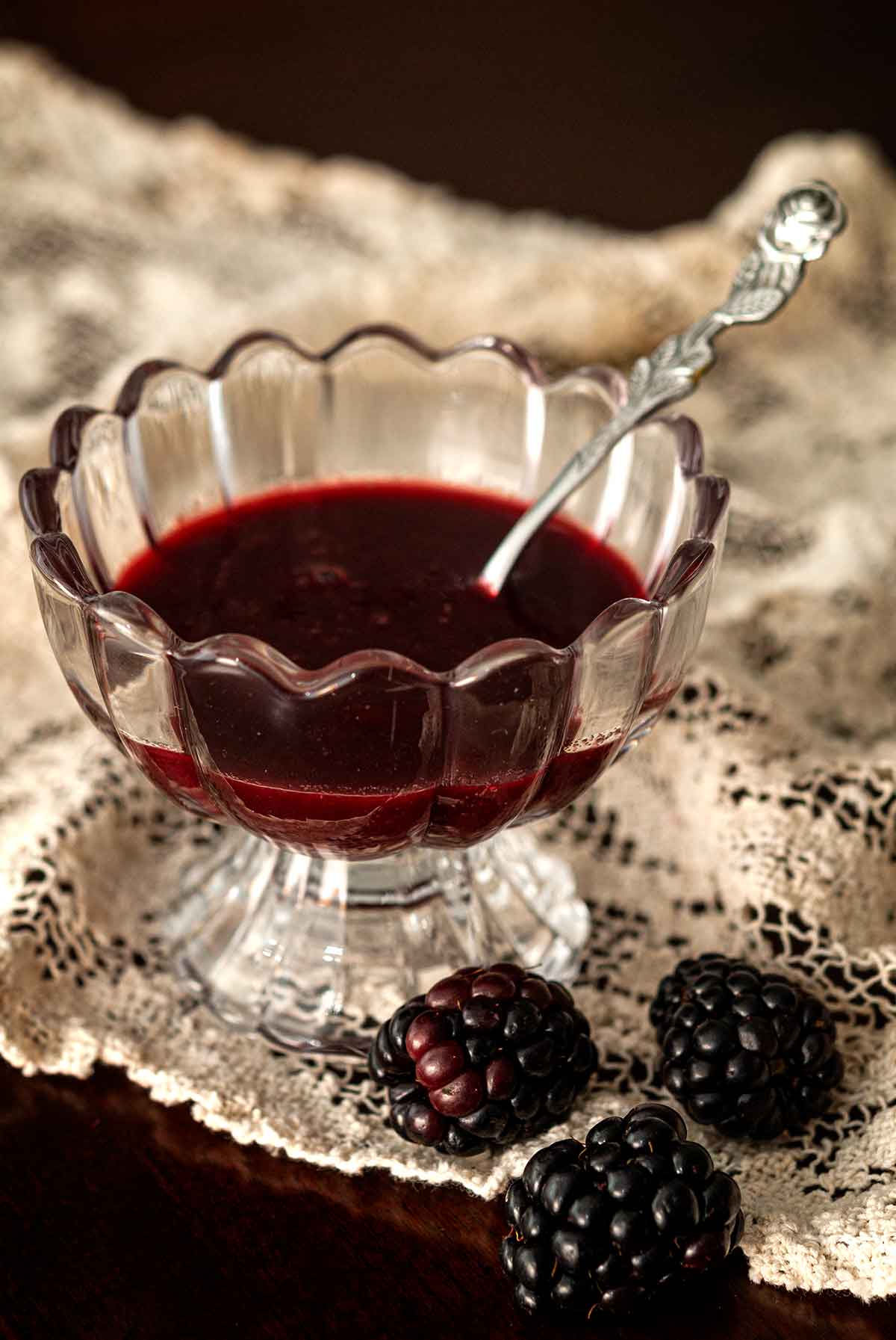 A decorative glass bowl of seedless blackberry sauce on lace beside 3 blackberries.