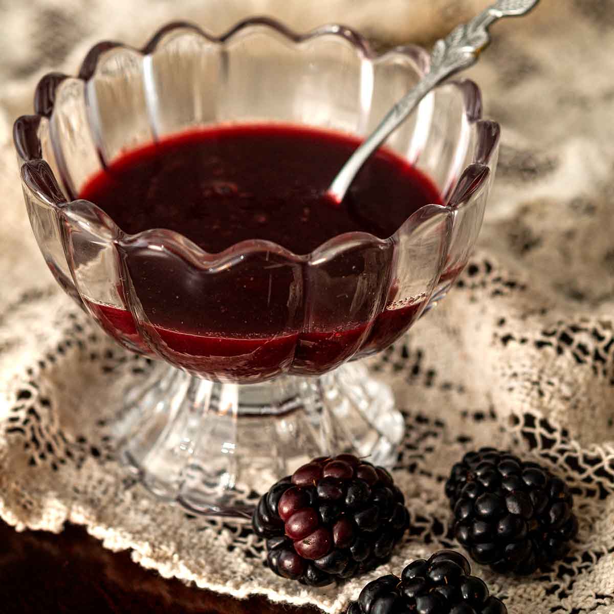 A decorative glass bowl of seedless blackberry sauce on lace beside 3 blackberries.