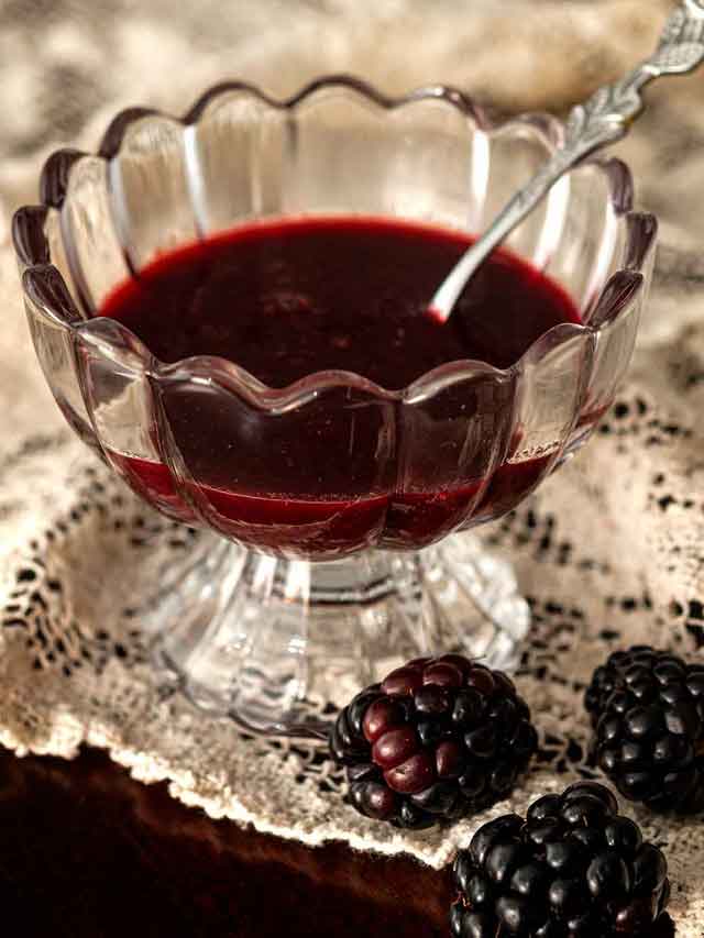 A decorative glass bowl of seedless blackberry sauce on lace beside 3 blackberries.