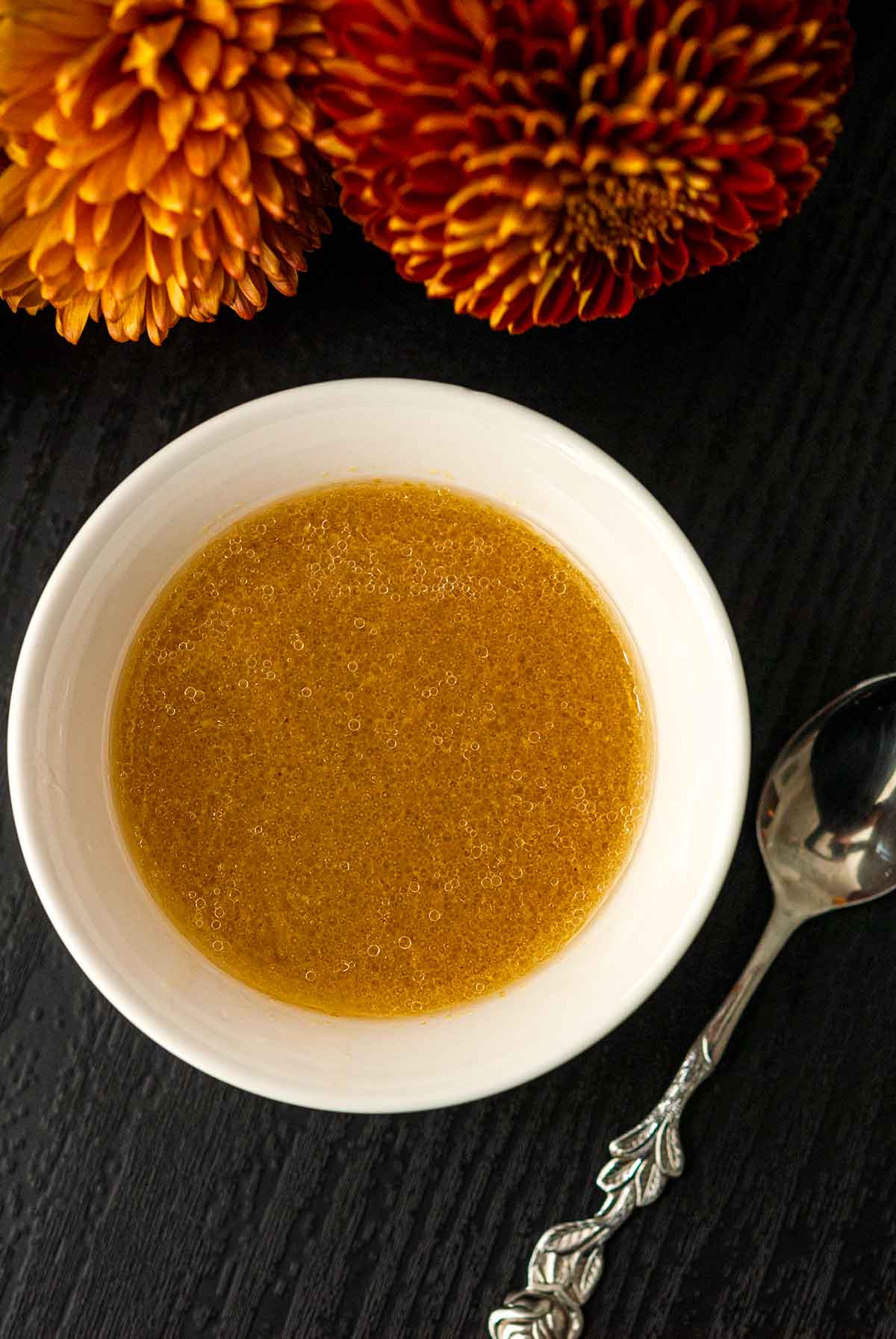 A bowl of apple cider vinaigrette on a table in front of flowers, beside a small spoon.