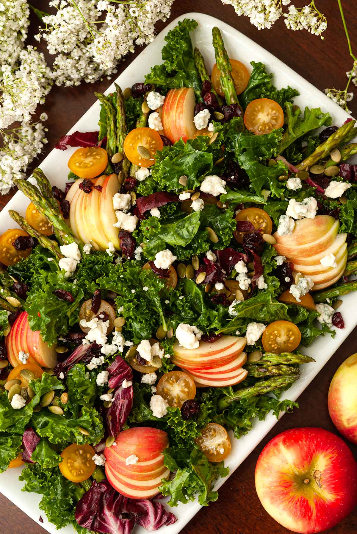 The top of an apple salad, sprinkled with cheese, beside 2 apples and flowers.