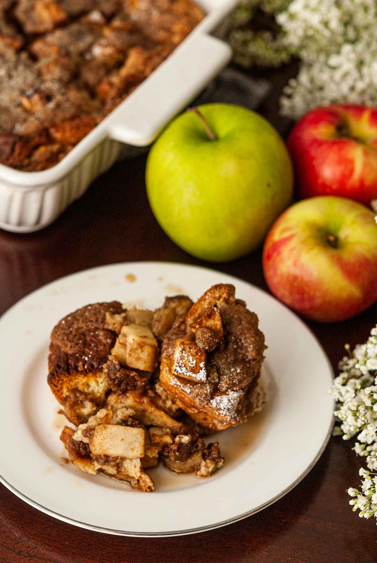 A sliced of french toast on a plate in front of a baking dish, beside 3 apples.