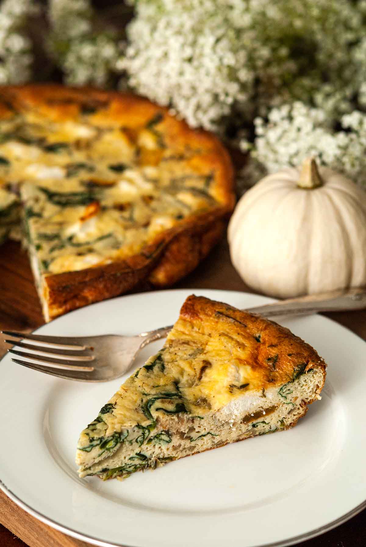A slice of quiche on a plate with a fork, in front of flowers, a pumpkin gourd and quiche.