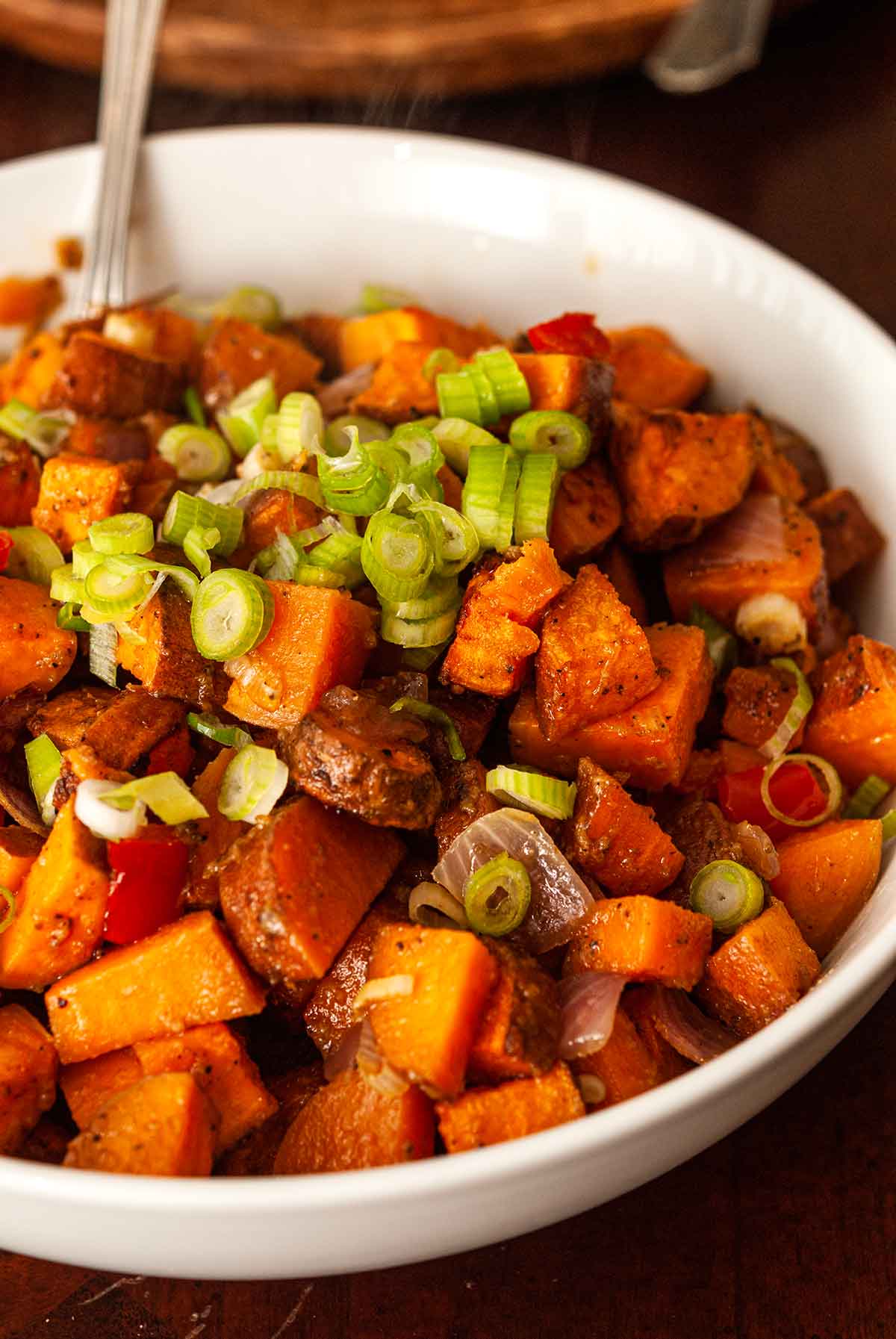 A bowl of sweet potato hash on a table, sprinkled with scallions with a spoon.