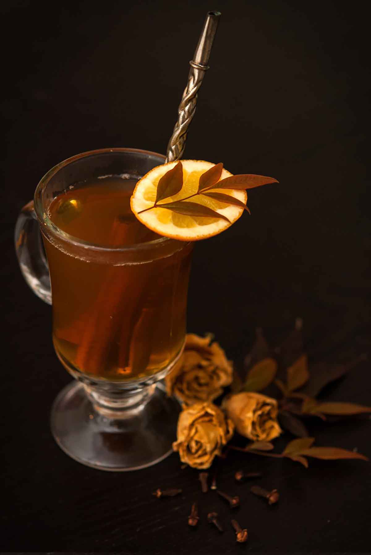 A hot toddy on a table with an orange and leaf garnish with a few dry roses.