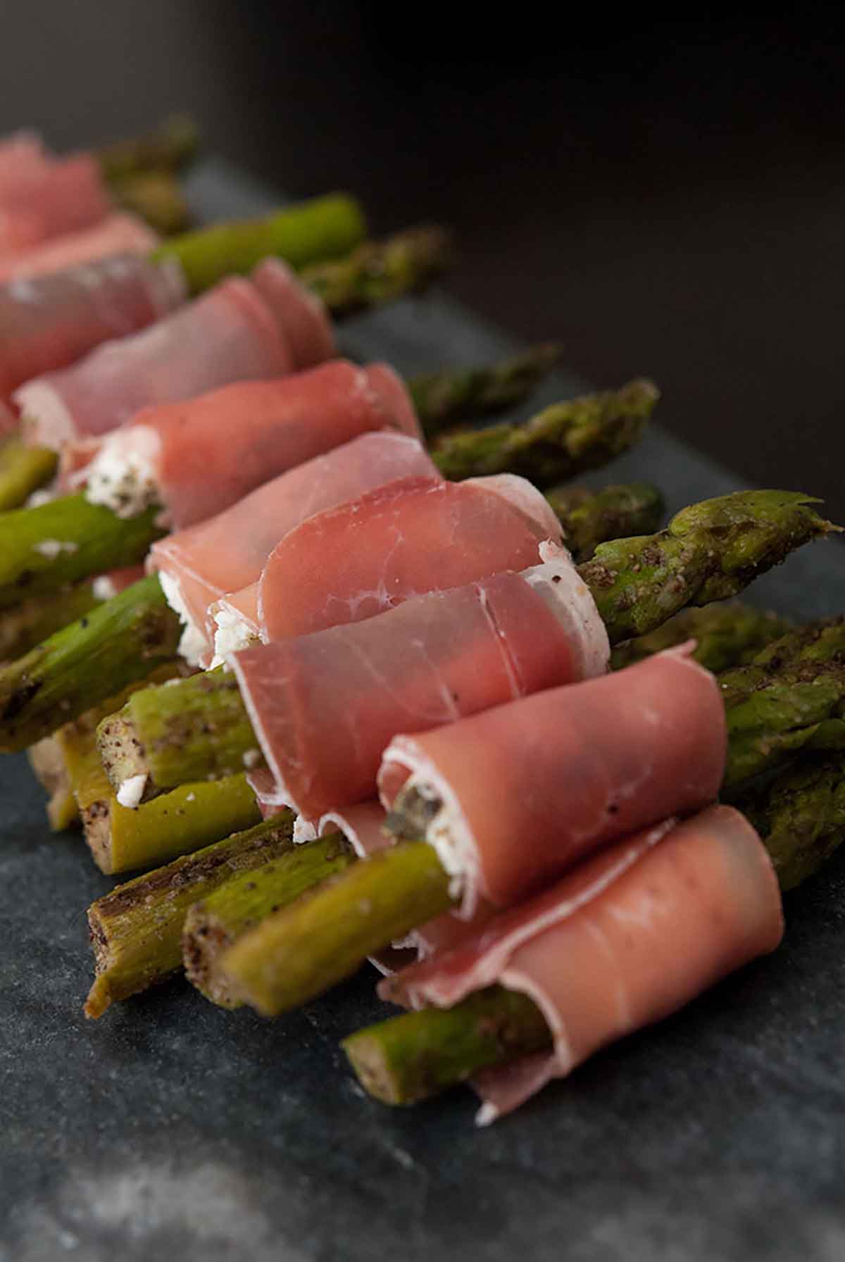 A pile of prosciutto-wrapped asparagus on a marble plate in a row.