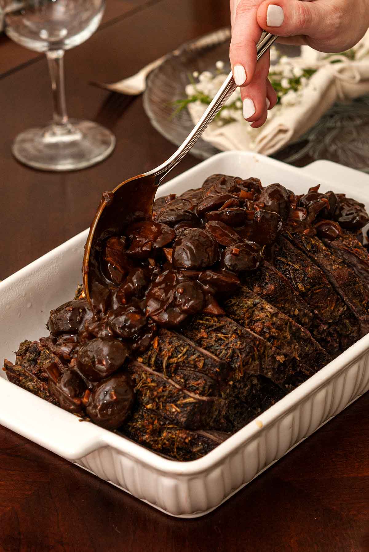 Top round roast beef in a tray with mushroom sauce being poured on with as spoon on a table.