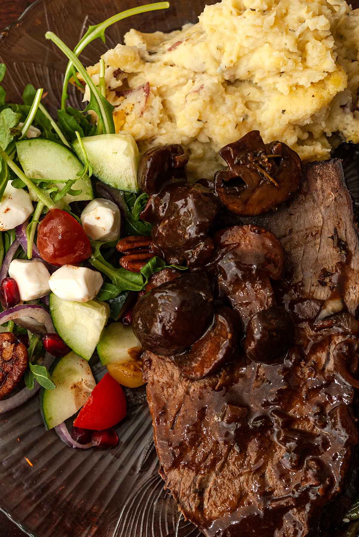 A slice of roast beef with mushrooms sauce, salad and mashed potatoes on a plate.