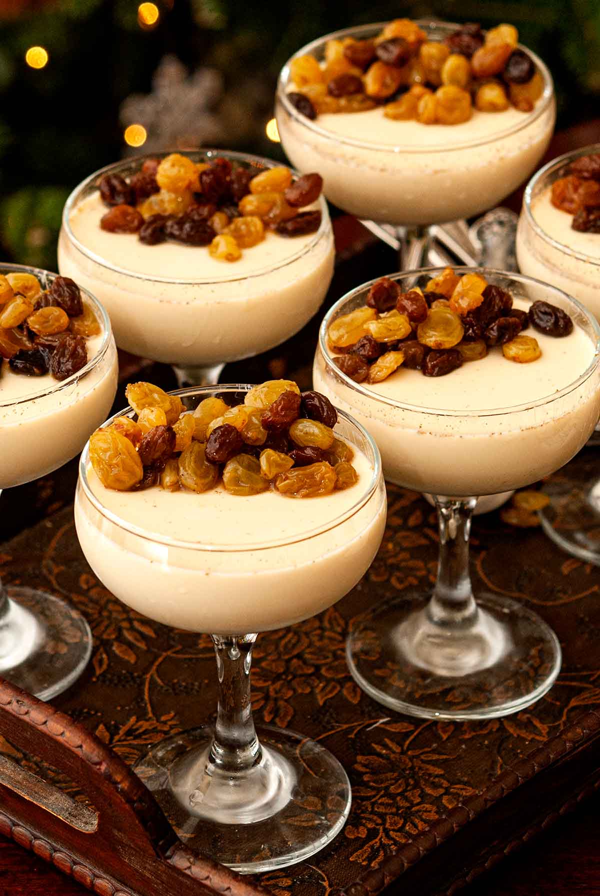 6 chai-spiced eggnog panna cottas with raisins on a serving tray in front of a Christmas tree.