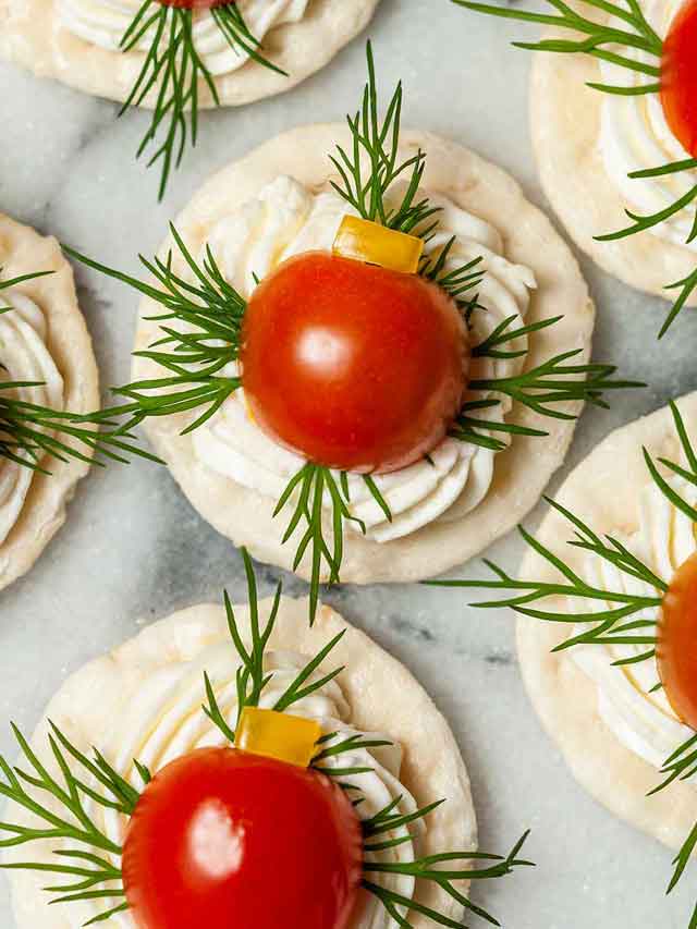 4 appetizers with cheese, dill, tomato and pepper on a cracker on marble.