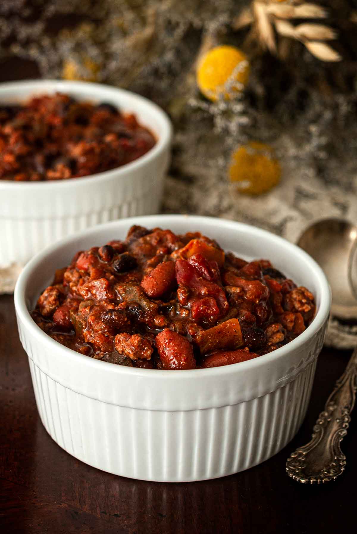 2 bowls of chili on a table beside dry flowers, spoons and a lace tablecloth.