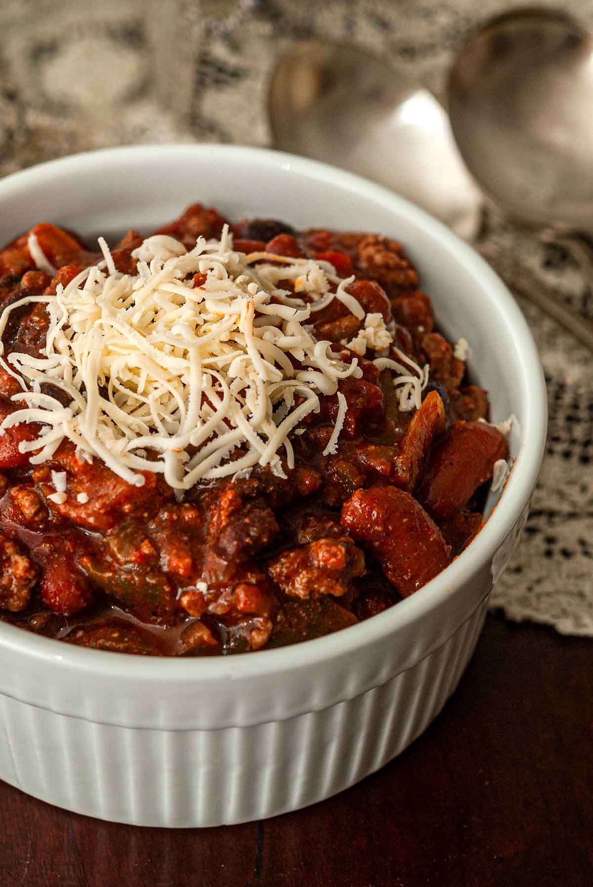 A bowl of chili on a table beside a lace table cloth, topped with cheese.