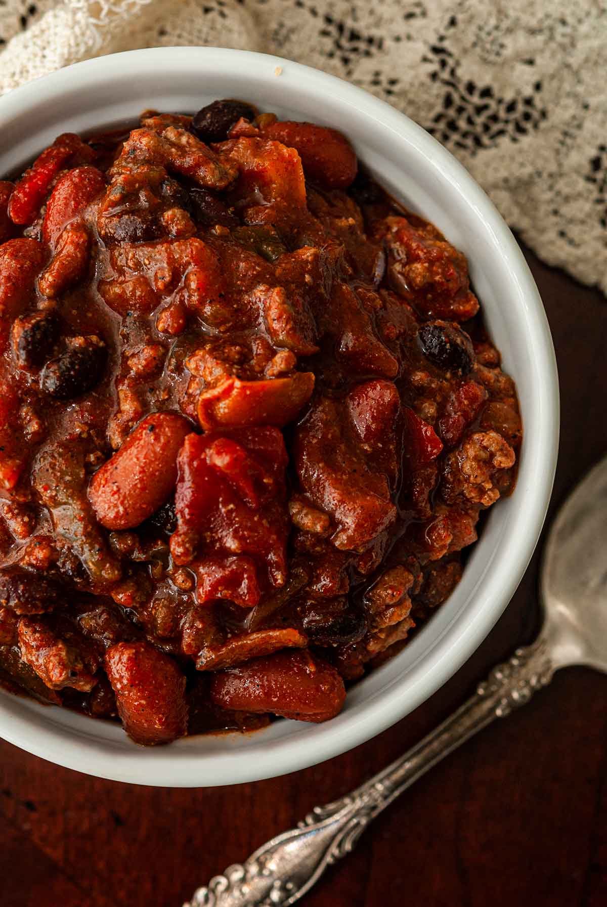 A bowl of chili beside a spoon and a lace tablecloth.