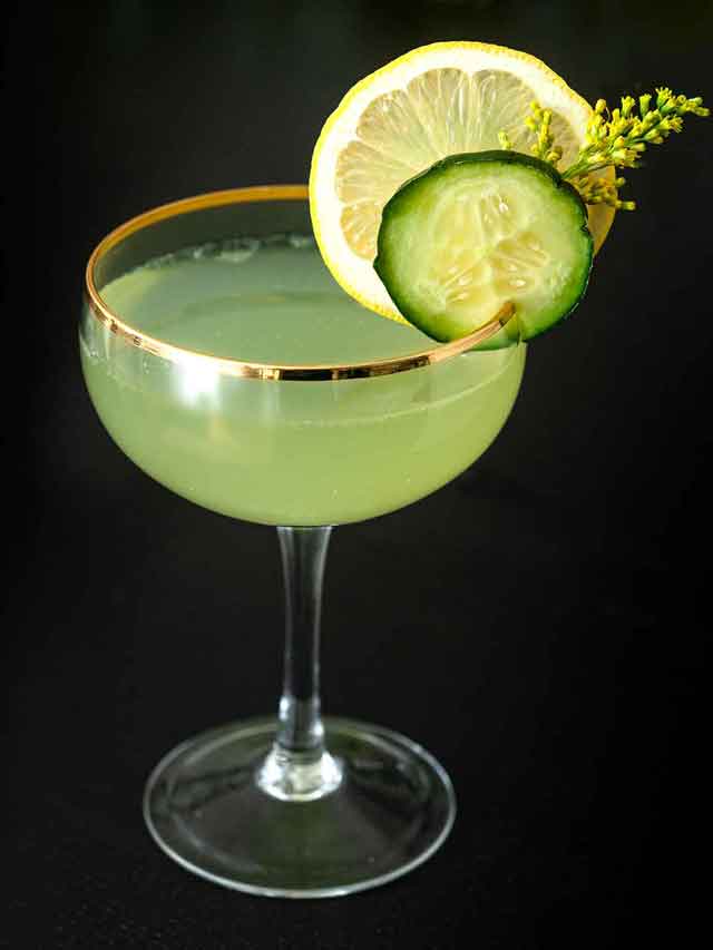 A cocktail in a coup glass, garnished with a cucumber slice, lemon, and wispy flower on a table.