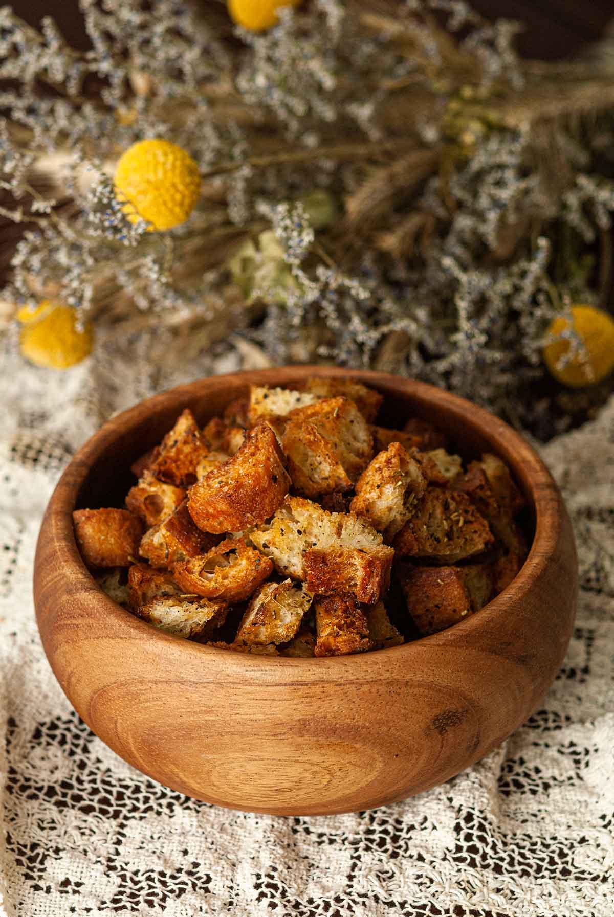 A bowl of Italian croutons on a lace table cloth in front of dry flowers.