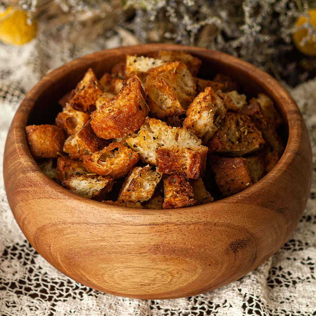 A bowl of Italian croutons on a lace table cloth in front of flowers.