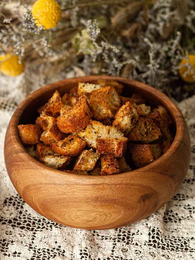 A bowl of Italian croutons on a lace table cloth in front of flowers.