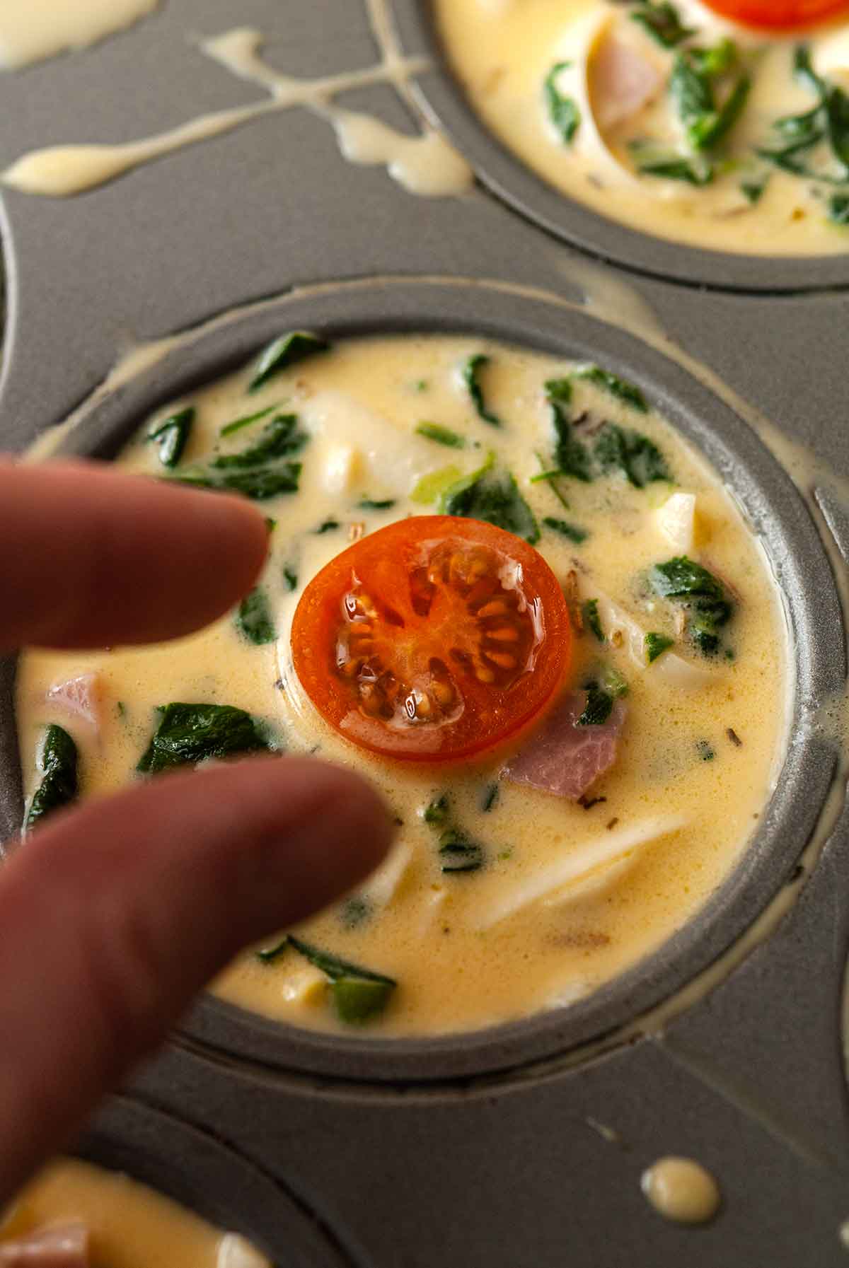 Fingers placing a tomato on an uncooked quiche in a tin.