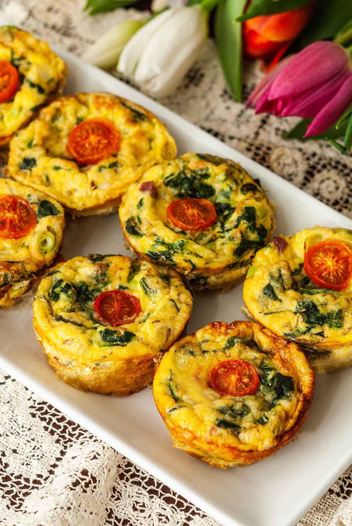 7 mini-quiches on a plate topped with tomatoes beside tulips on a table.