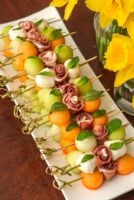 A long plate of melon bamboo skewers on a table beside daffodils in glass vases..