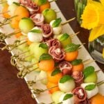A long plate of melon bamboo skewers on a table beside daffodils in glass vases..