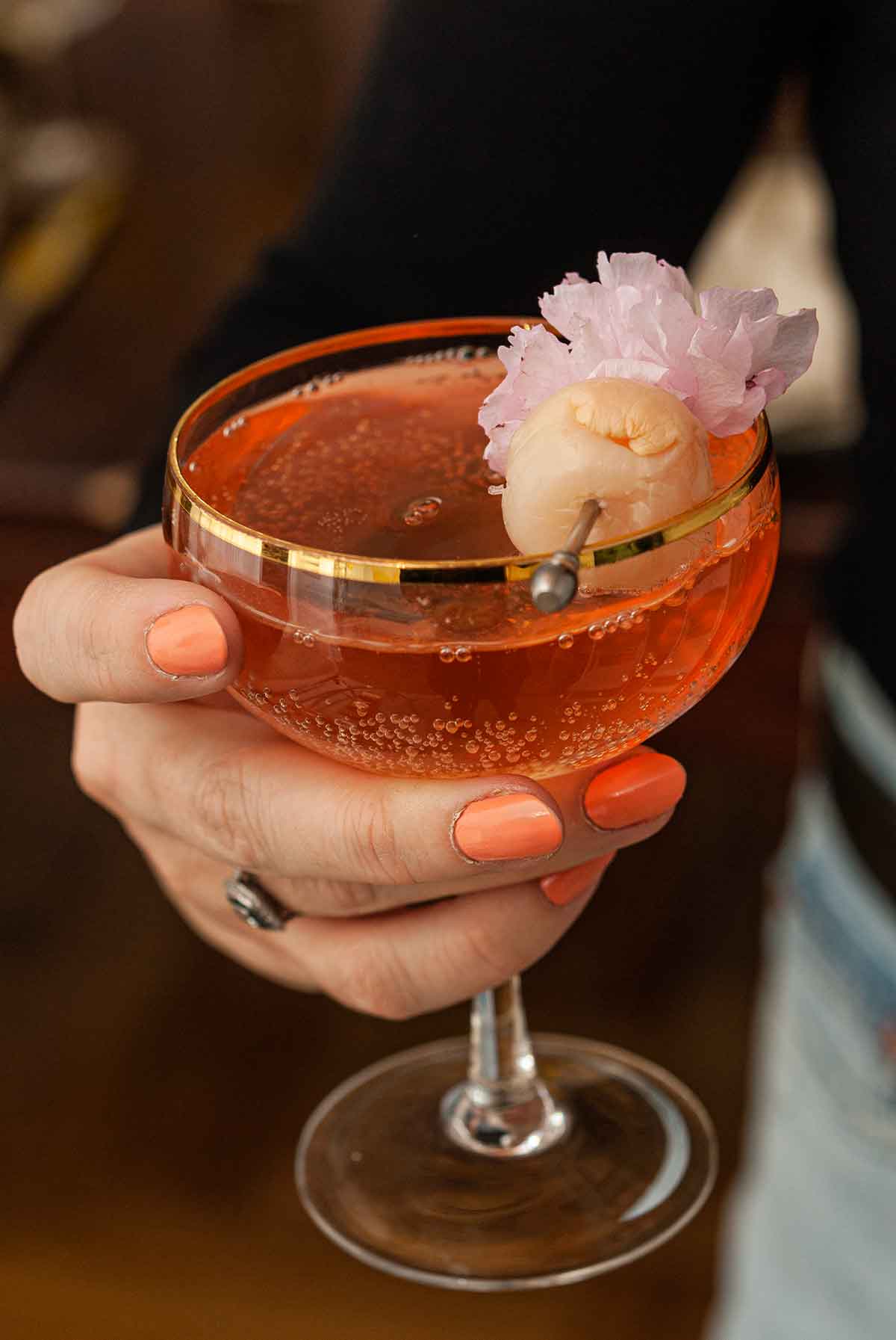 A women's hand holding a cocktail garnished with a lychee and cherry blossom.