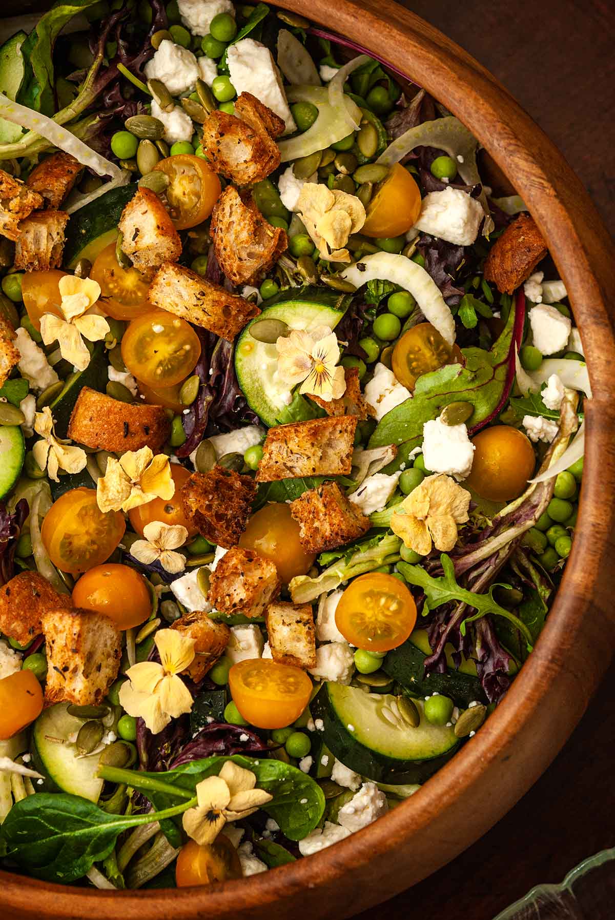 A salad with vegetables, cheese, croutons and flowers in a wooden bowl. 