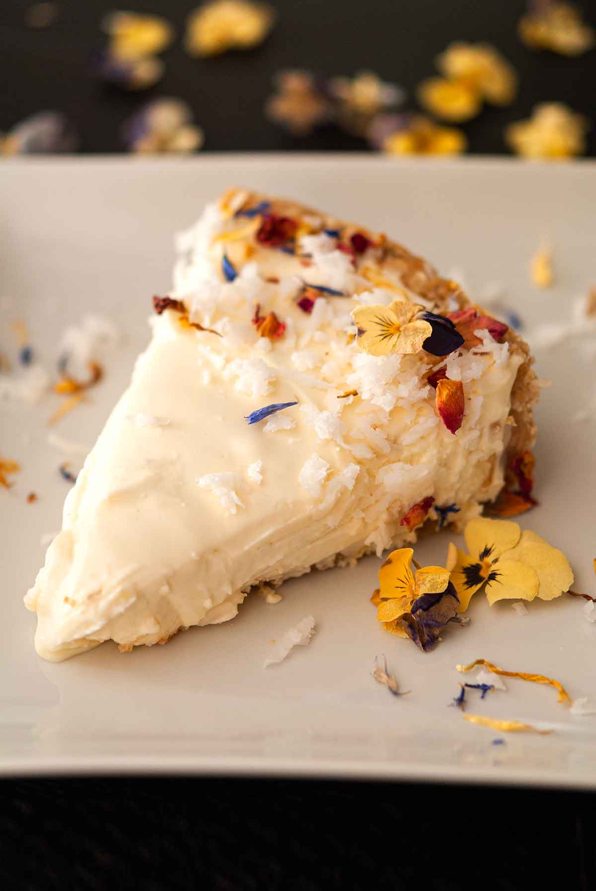 A coconut cheesecake slice garnished with coconut and flowers on a plate with pansies.