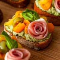 A pesto crostini with a prosciutto rose on a wooden board.