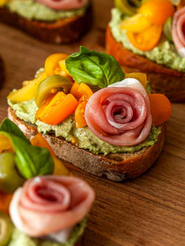 A pesto crostini with a prosciutto rose on a wooden board.