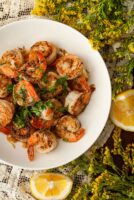 A bowl of lemon butter shrimp beside flowers and a lemon.