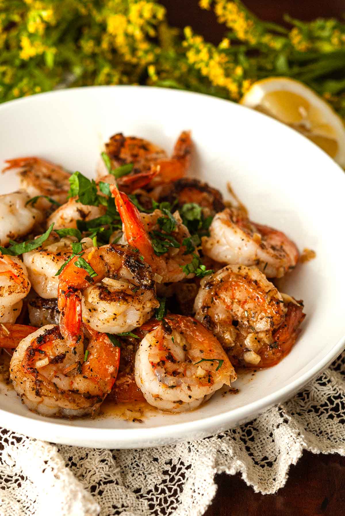 A bowl of lemon butter shrimp on a lace tablecloth beside flowers and a lemon.