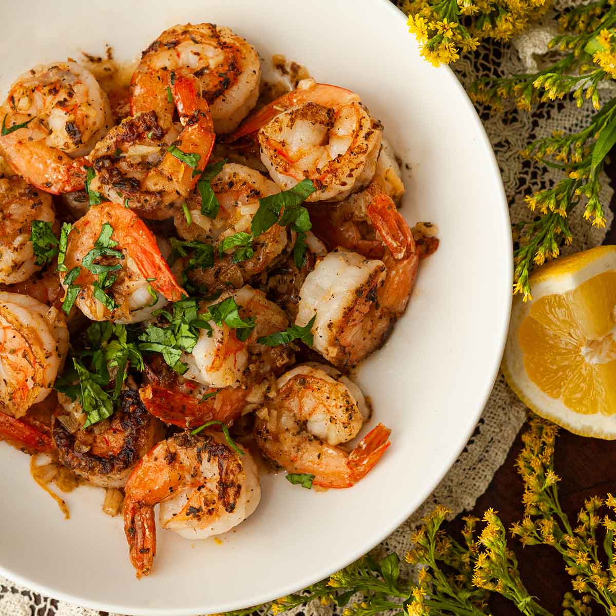 A bowl of lemon butter shrimp beside flowers and a lemon.