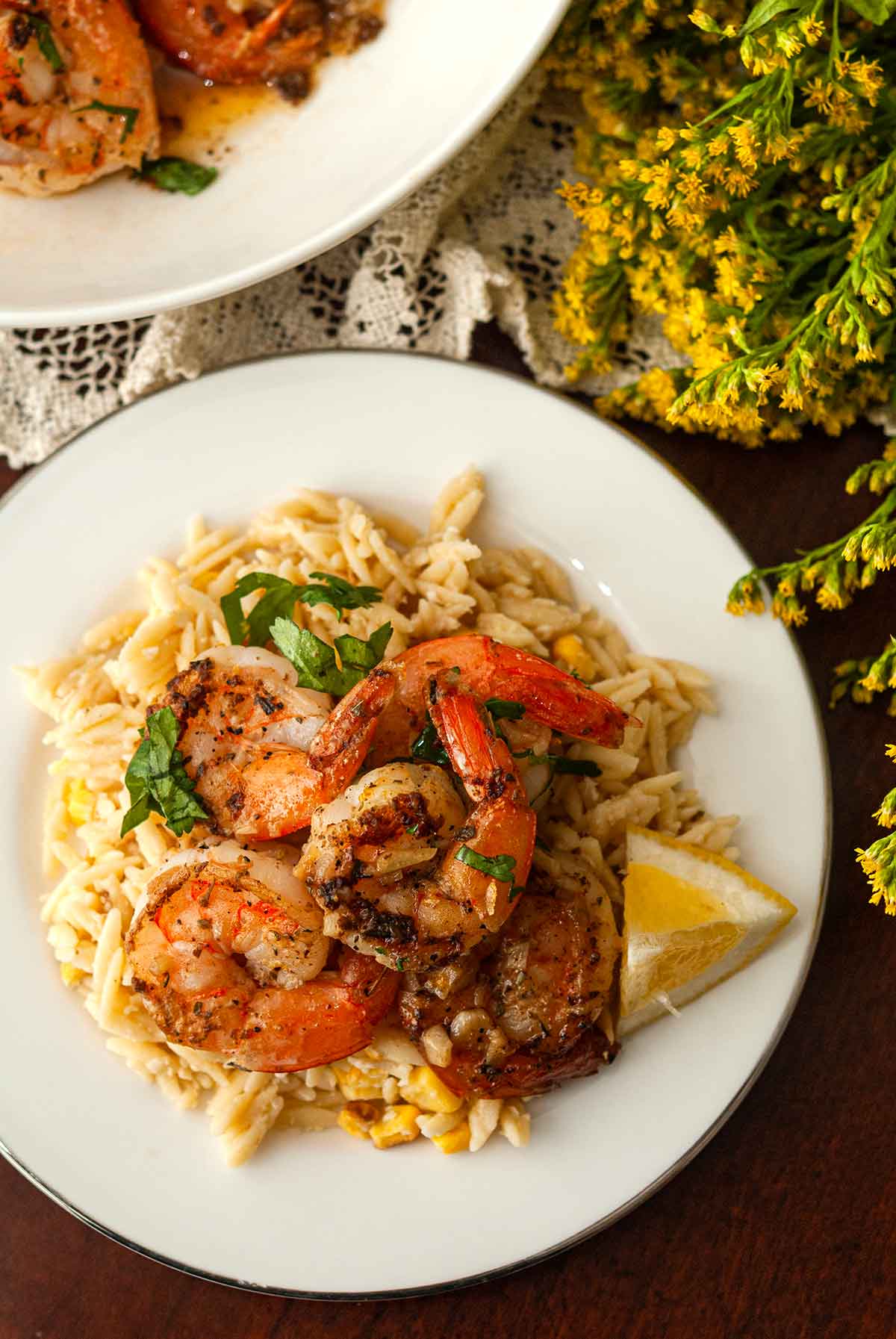 A plate of lemon shrimp and lemon orzo on a table beside flowers.